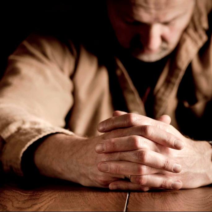 a white man with his hands clasped as if praying, sitting at table sad, thoughtful, depressed
