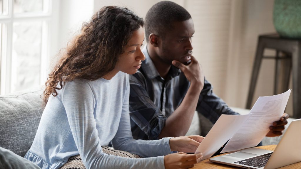 a young Black woman and young Black man sitting together on a couch looking seriously at information on a computer and reading paper documents