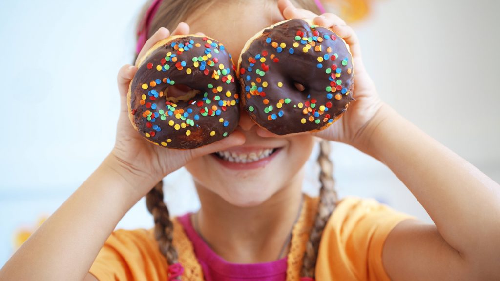 a young white girl with blonde braids holding sprinkle covered donuts over her eyes and smiling