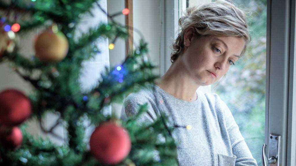 a young white woman looking sad and depressed, leaning against a glass door, with a decorated Christmas tree in the foreground