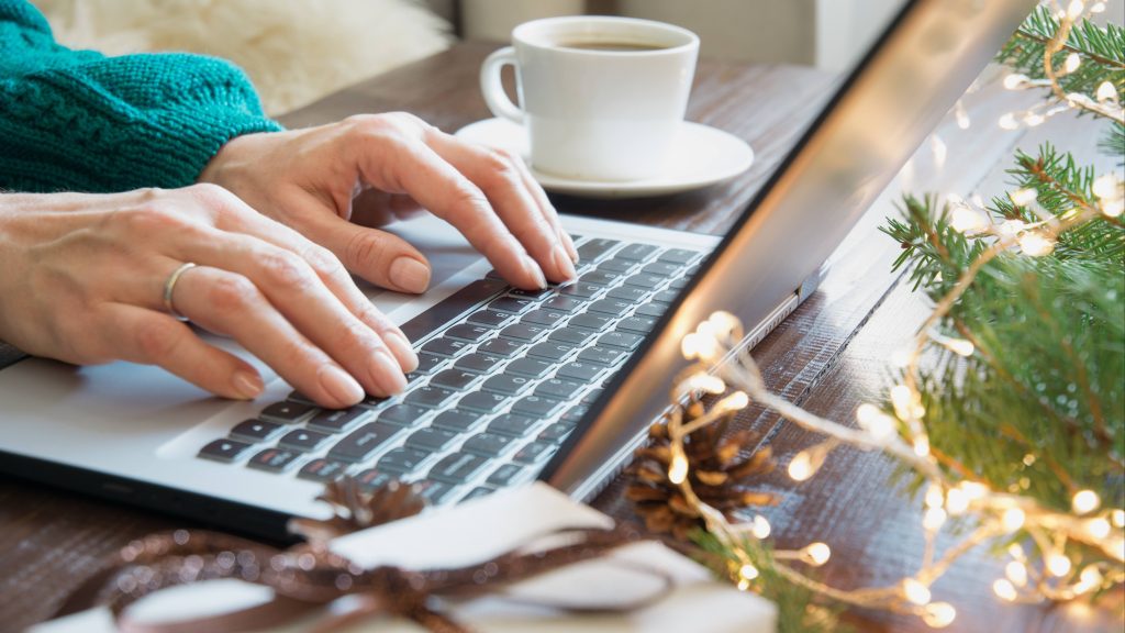closeup of a white woman's hands typing on a computer laptop with a cup of coffee nearby and a small Christmas tree with tinsel on the table