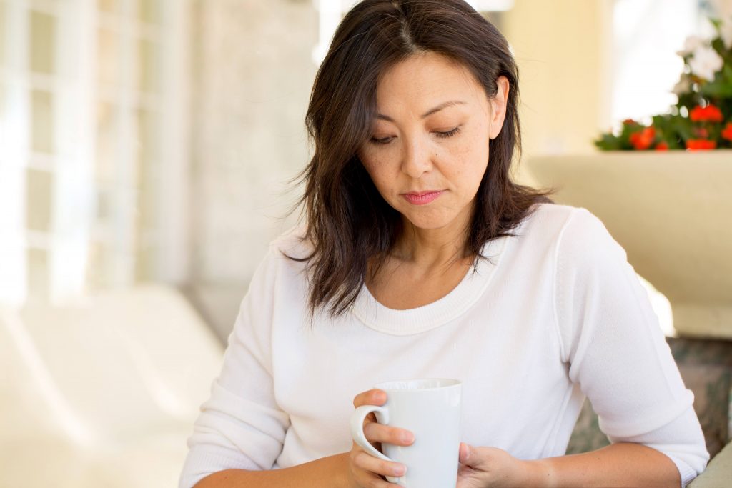 a middle aged woman with light brown skin tone holding a coffee mug and looking sad, worried, depressed