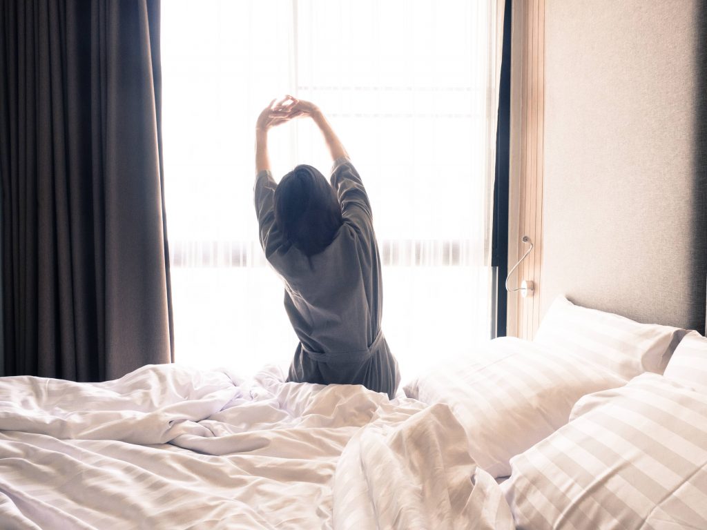 a white woman sitting on the edge of a bed in a robe, looking out a window and stretching her arms up in the air for morning exercise