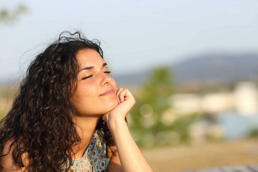 a young woman, perhaps older teenager, with light brown skin and long dark curly hair, smiling with her eyes closed and looking toward the sunshine
