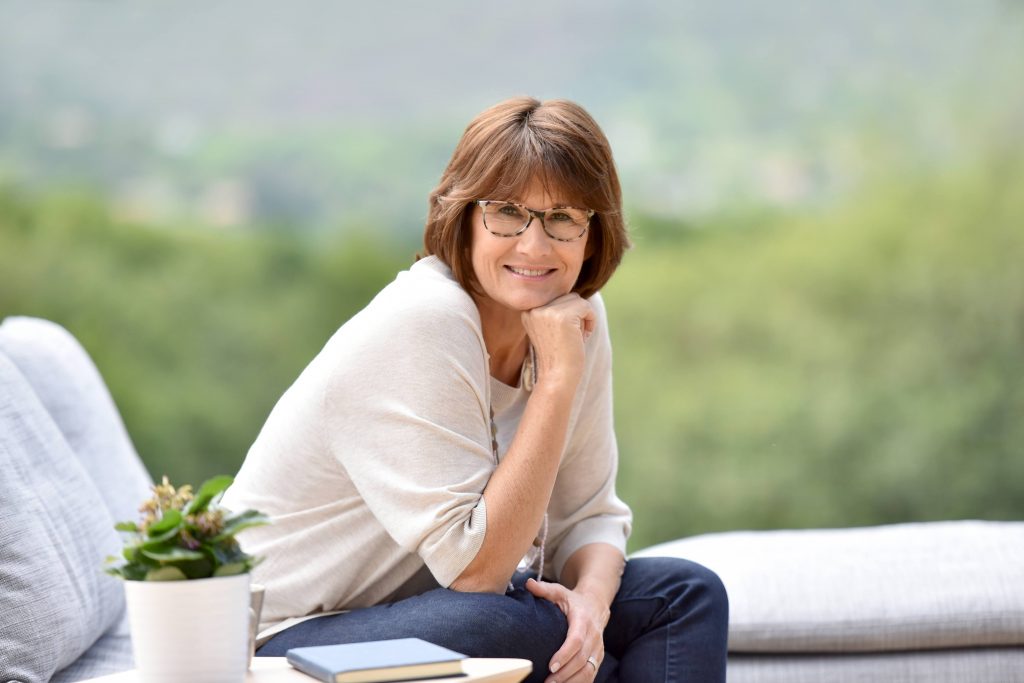 a white woman with short brown hair and glasses, sitting on an outside couch and smiling