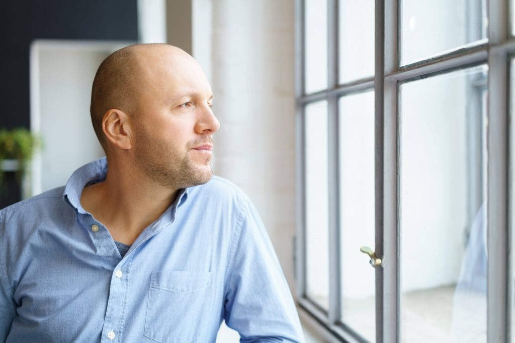 a white man in a blue shirt, looking serious, worried and thoughtful while standing at a window looking out