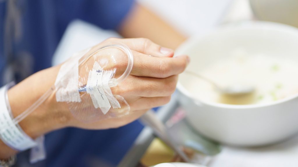 closeup of a white person's hand with an IV, a hospital patient, holding a spoon and eating food from a bowl