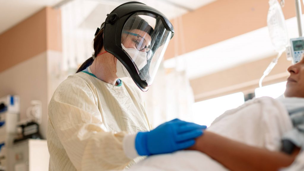 a Mayo Clinic health care worker, a white woman, perhaps a physician or nurse in full COVID-19 PPE attending to a patient in bed