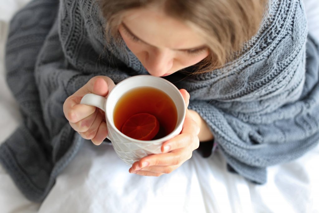 a young white woman, sick with a cold, wrapped in a gray knitted blanket holding a warm cup of tea