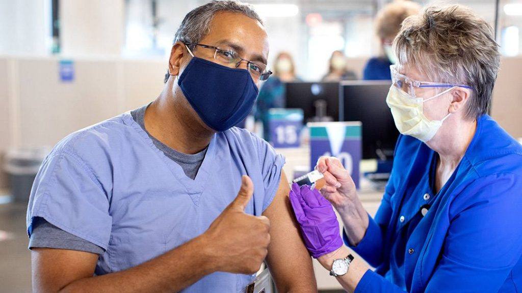 a Black man, Mayo Clinic medical worker in blue scrubs and wearing a mask while receiving a COVID-19 vaccine from a fellow Mayo employee, a white woman wearing PPE
