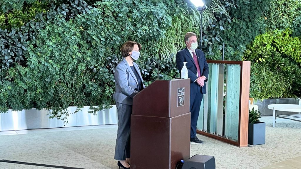 U.S. Senator Amy Klobuchar wearing a mask standing at a podium speaking at news conference with Dr. Andrew Badley, wearing a mask, standing at a distance