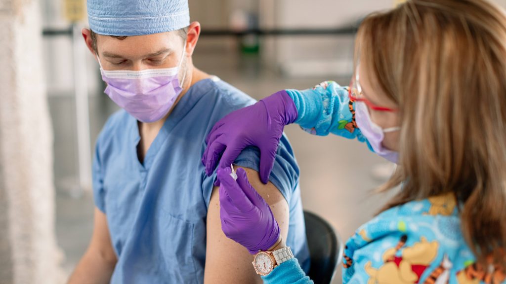 a Mayo Clinic employee, a white man, in blue scrubs and wearing a face mask receiving the COVID-19 vaccine from a white woman wearing purple PPE mask and gloves