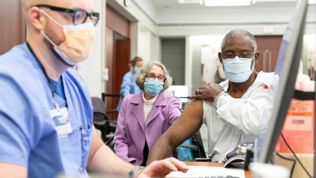 a Black adult man wearing PPE rolling up the sleeve of his white shirt for his COVID-19 vaccine