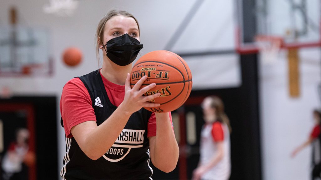 a high school basketball player, a white girl, wearing a uniform and face mask, getting ready to shoot a basketball