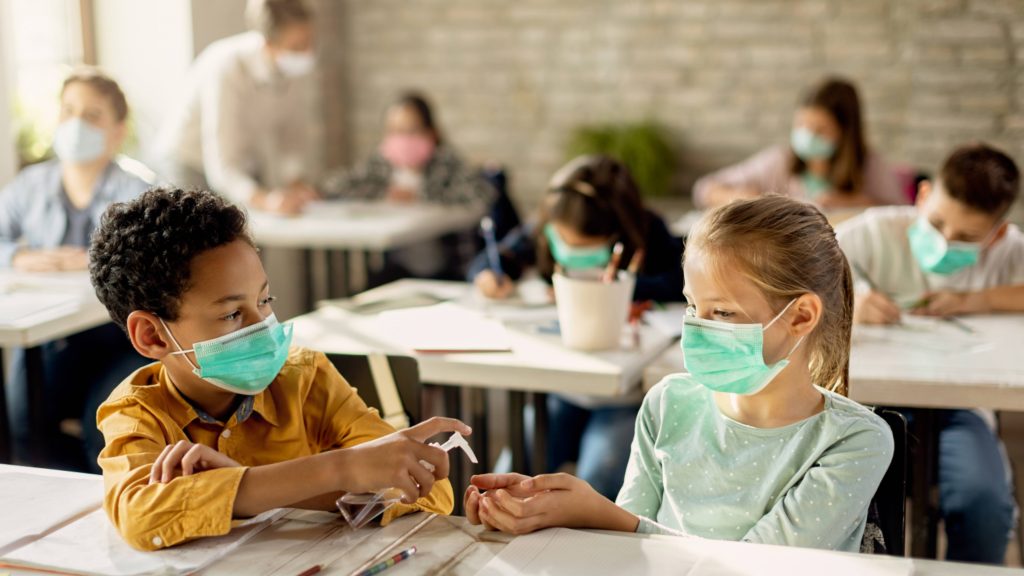 Dos pequeños, un niño negro y una niña blanca, con sendas mascarillas están sentados en sus pupitres en el aula y comparten el desinfectante de manos 