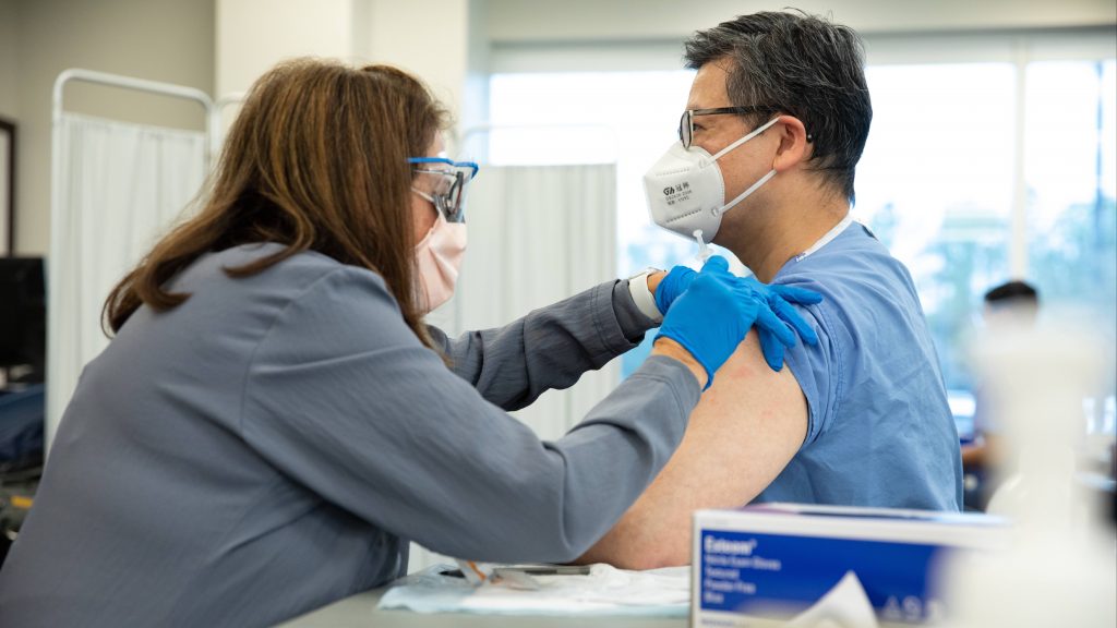 Mayo Clinic medical staff person (a white woman) wearing PPE administering a COVID-19 vaccine to a a man, perhaps Asian American, also wearing PPE