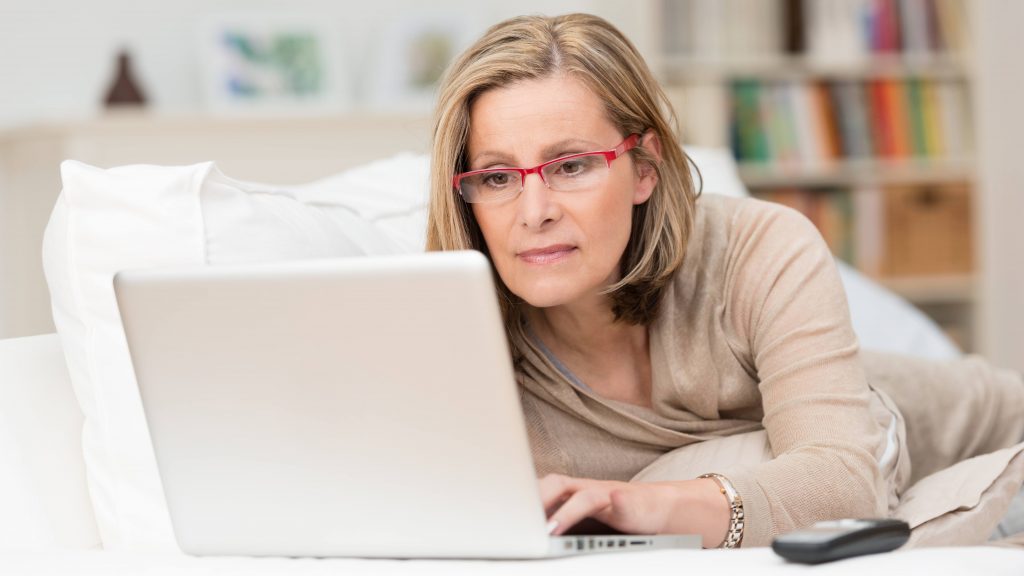 a middle-aged white woman wearing glasses and working on a laptop computer on her bed