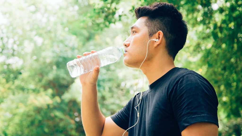 a young Asian American man outside exercising, wearing ear buds for music and drinking a bottle of water