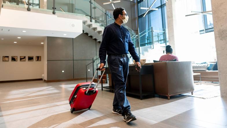 a man, perhaps Latino, wearing a mask walking through a hotel lobby with a traveling suitcase
