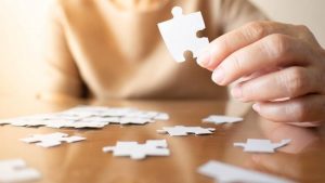 a medium shot of a white woman sitting at a table with a jigsaw puzzle holding a puzzle piece