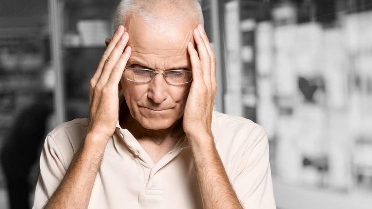 an elderly white man with grey hair looking sad, worried, forgetful holding his head with his hands
