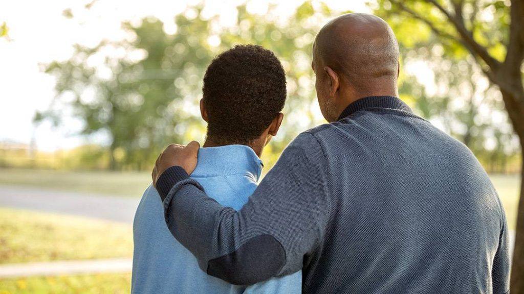 a Black man, perhaps a father, with his arm on the shoulder of a young Black boy in support and comfort