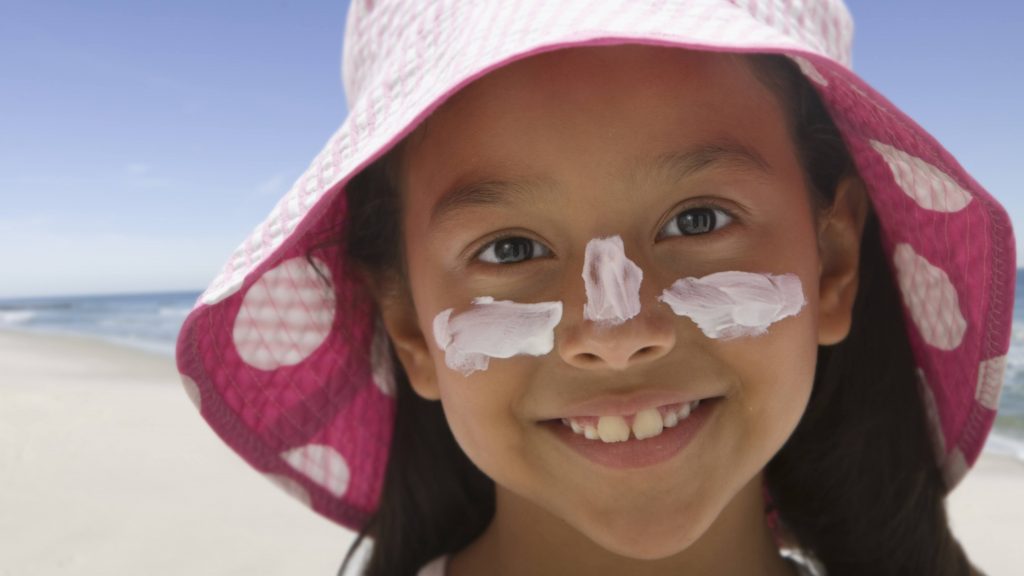 a little girl, perhaps Latina, at the beach smiling and wearing a sun hat, sunscreen
