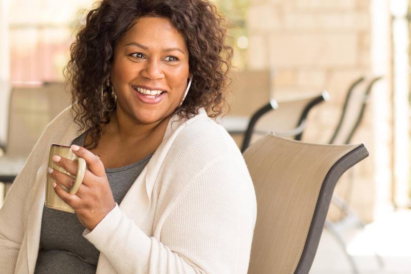 a middle aged Black woman holding perhaps a cup of coffee, sitting by a window smiling and looking happy
