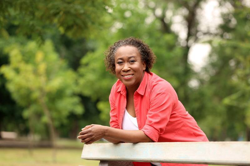 a middle aged Black woman outside, leaning on a fence, smiling and looking happy