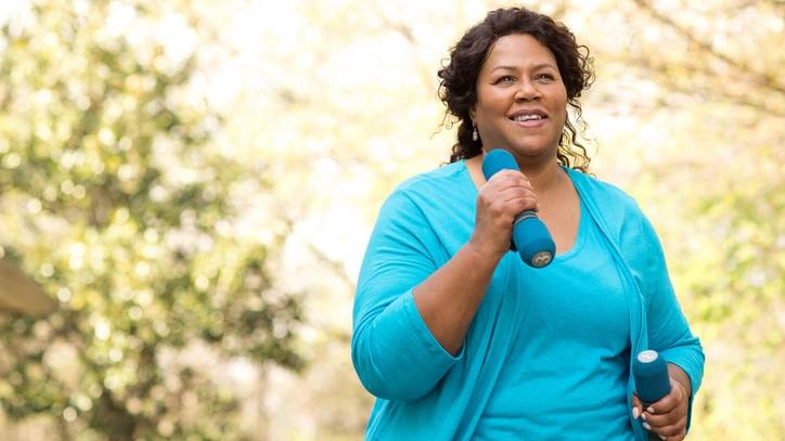 a middle aged Black woman outside, smiling while exercising, walking and carrying hand weights