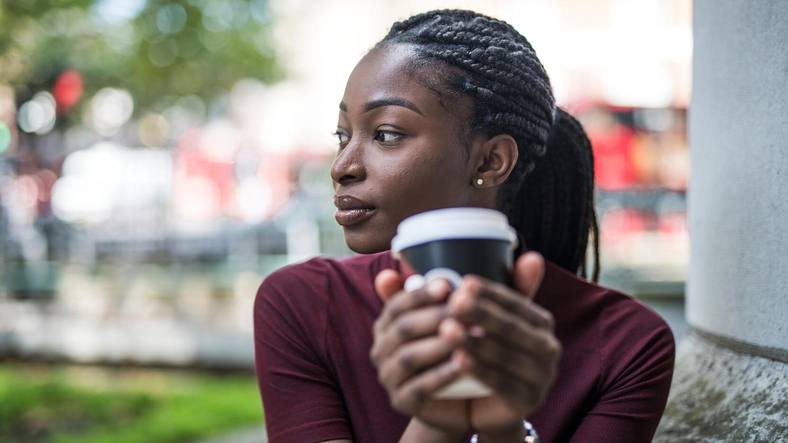 a young Black woman sitting outside holding a cup of coffee, looking serious and thoughtful