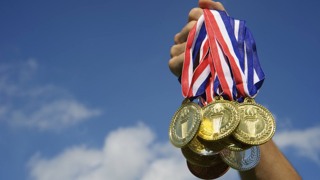 closeup of a person's arm holding up a handful of Olympic gold medals with ribbons against a blue sky