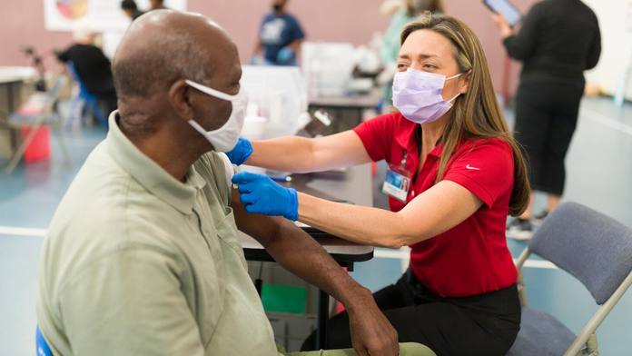 a Mayo Clinic medical staff person, a white woman, administering a COVID-19 vaccine to a Black man, both wearing masks in a vaccine clinic