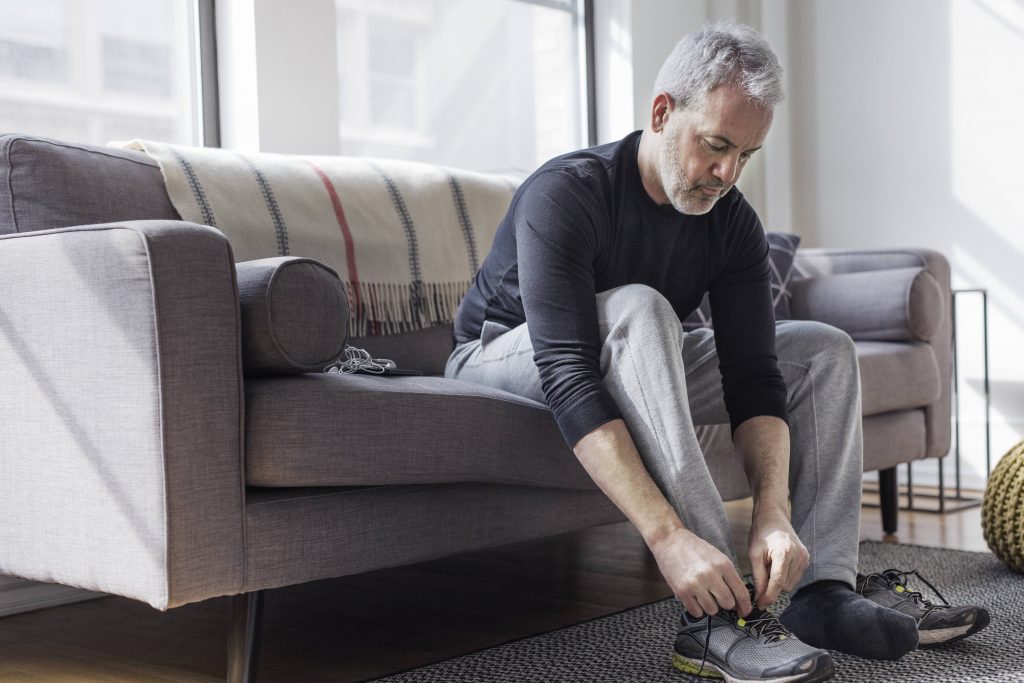 a middle aged white man in sweatpants sitting on a couch and tying exercise shoes