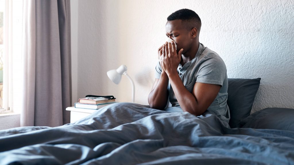a young Black man sick in bed, coughing and blowing his nose