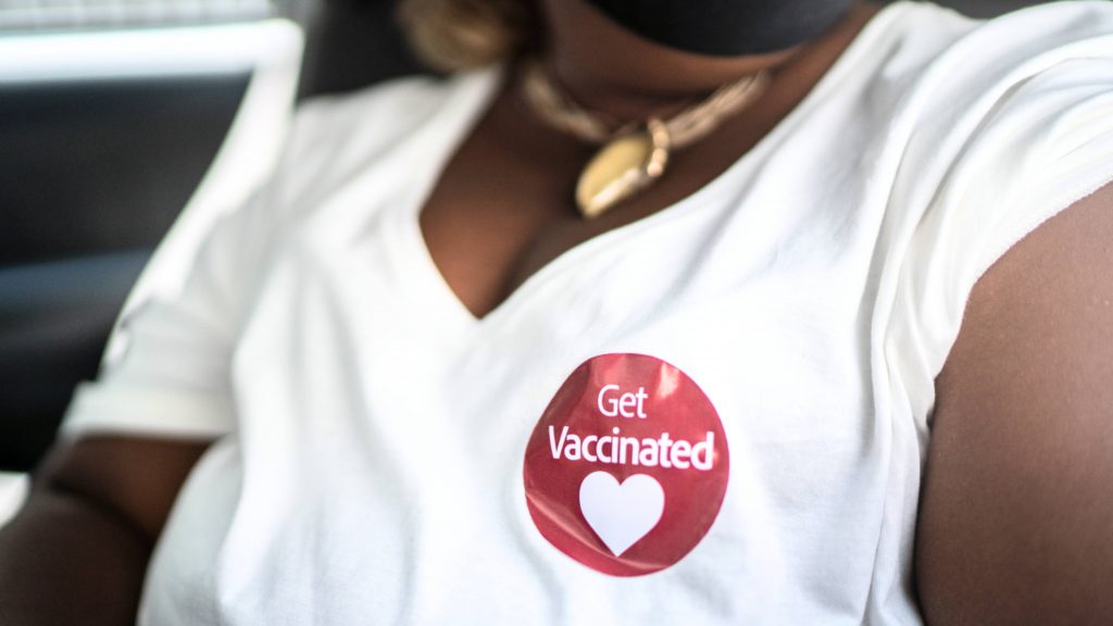 a young Black woman in a white t-shirt with a sticker saying 'Get vaccinated'