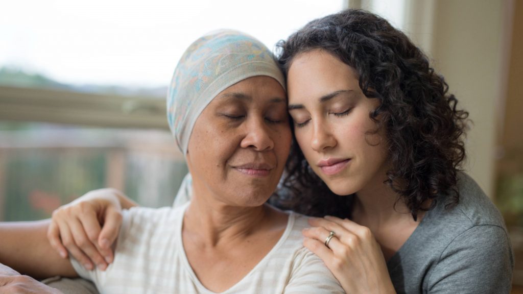 a young woman, perhaps Latina or Asian, sitting by a window comforting an older woman wearing a headwrap, perhaps a relative with cancer