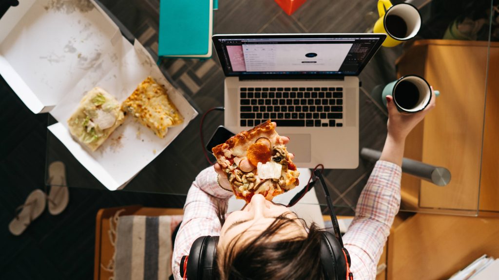 a young woman sitting at a home computer eating pizza with two cups of coffee nearby, maybe stressed with work