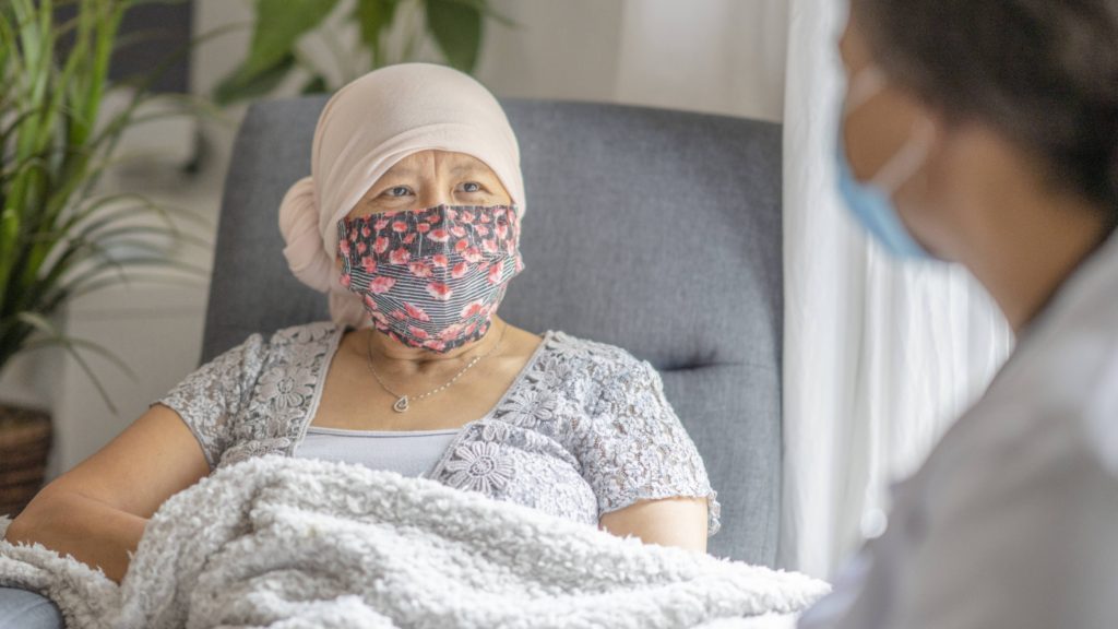 an older Asian woman, maybe patient with cancer, wearing a headscarf and mask, sitting in room with nurse or doctor