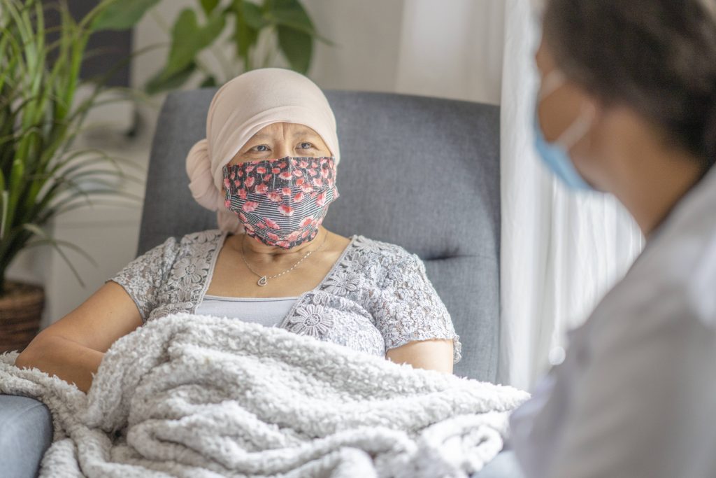 an older Asian woman, maybe patient with cancer, wearing a headscarf and mask, sitting in room with nurse or doctor