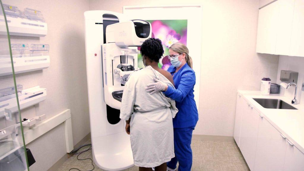 A Black woman in a patient gown being prepared by a medical assistant for a breast mammogram for possible dense breast