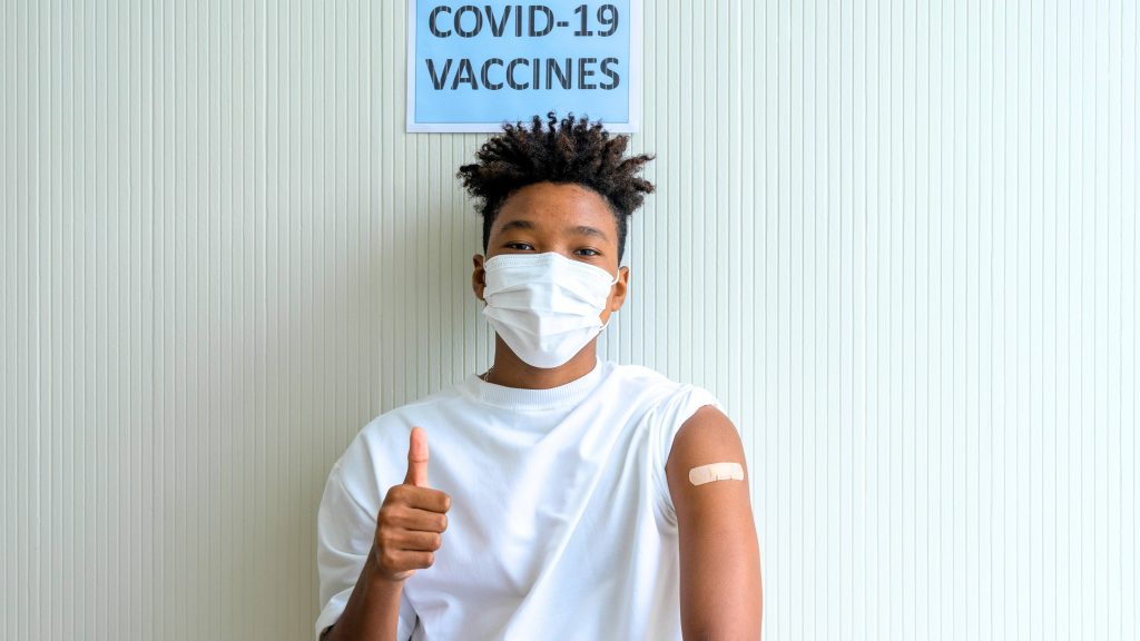 a young Black teenage boy with a bandaid on his arm, wearing a mask with his thumb up and standing by a sign that says COVID-19 vaccine