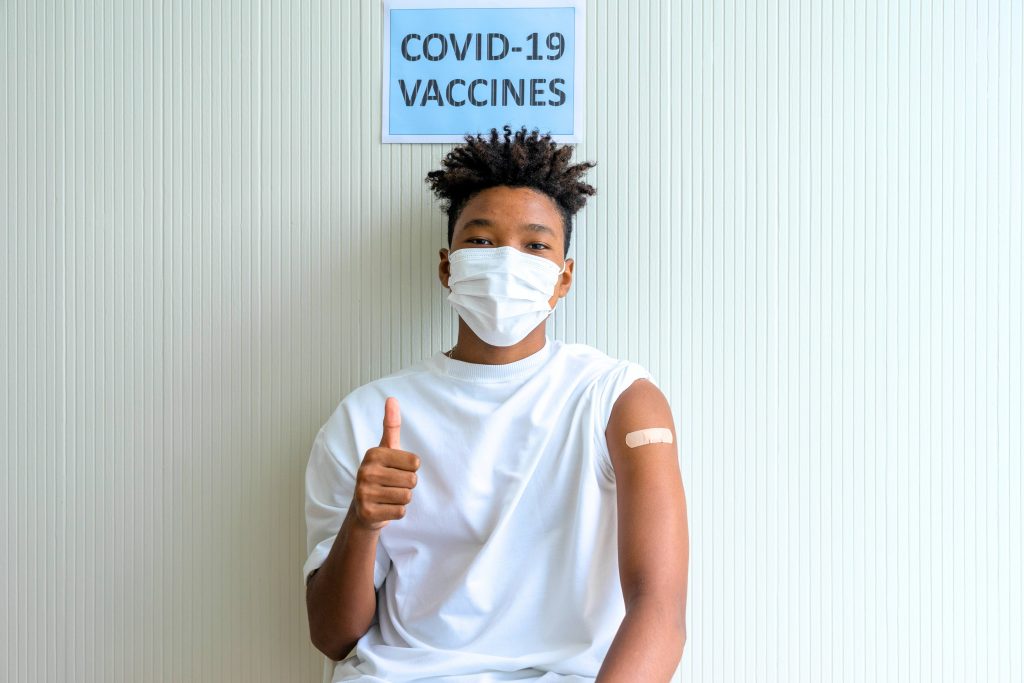a young Black teenage boy with a bandaid on his arm, wearing a mask with his thumb up and standing by a sign that says COVID-19 vaccine