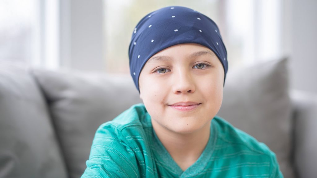 a young white child softly smiling, wearing a scarf wrap as if they have cancer and are receiving chemo therapy