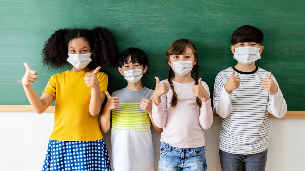 four school aged children wearing masks and standing near chalkboard with their thumbs up