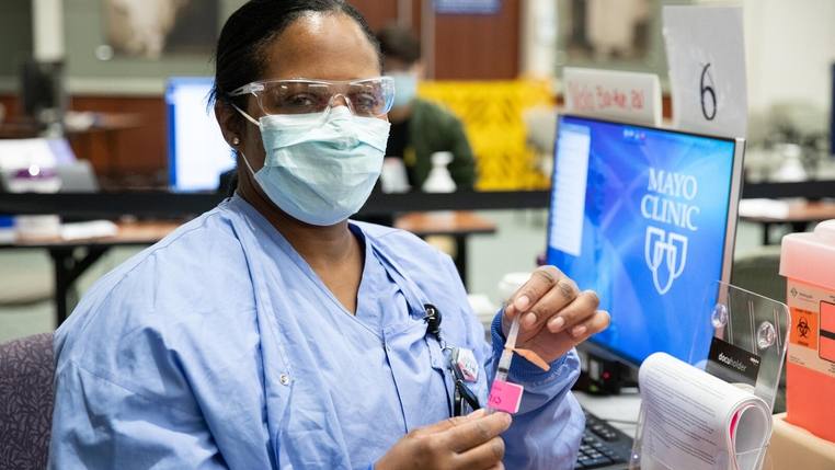 a Black woman, Mayo Clinic nurse or medical tech person, wearing a mask and holding a COVID-19 vaccine syringe