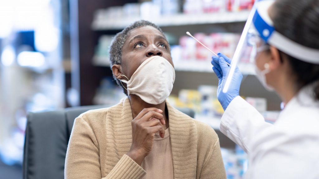 a middle-aged Black woman pulling down her mask to be swabbed and tested for COVID-19 by a pharmacist wearing PPE