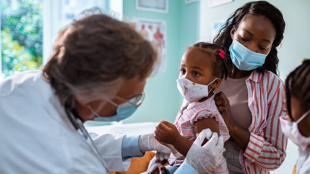 a Black woman, perhaps the mother, holding a child in doctor's office getting a vaccine, all wearing masks