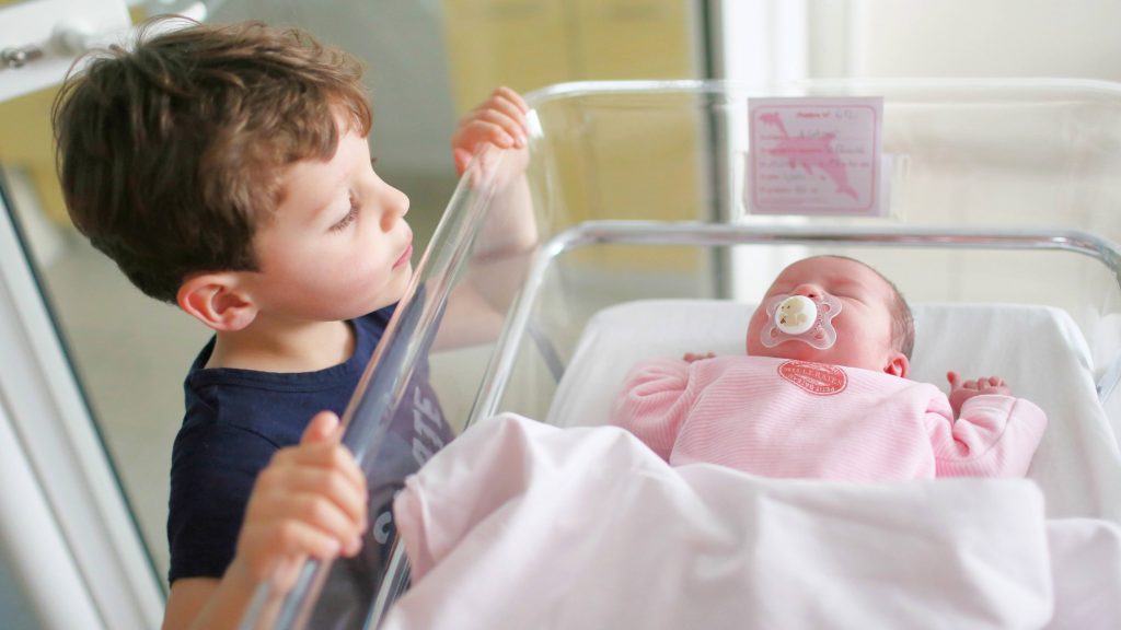 a little white boy peaking at his newborn infant sister in a hospital bassinet