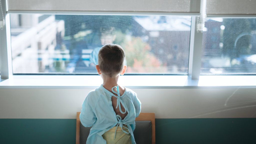 a sick, young boy in a hospital gown, maybe recoverying after surgery, looking out a window